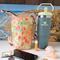 The back of an orange & yellow floral print water bottle bag is displayed. Beside the bag is a Stanley 30 ounce Flip Straw Tumbler.